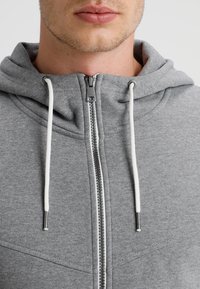 Primo piano della parte inferiore del volto di una persona che indossa una felpa grigia con zip e lacci bianchi su uno sfondo semplice.