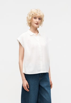 Νεαρό άτομο με κοντά σγουρά ξανθά μαλλιά που φοράει λευκό, φαρδύ, κοντομάνικο πουκάμισο και μπλε τζιν με φαρδιά πόδια.