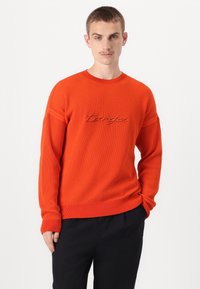 Maglione arancione a coste con scollatura rotonda e nome del brand ricamato sul petto. Maniche lunghe e finitura testurizzata.