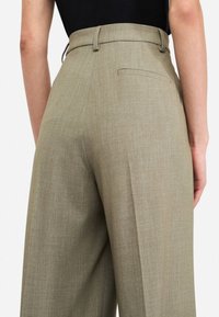Pantaloni beige su misura a vita alta con una tasca posteriore, fronte plissettato e tessuto a trama liscia. Dettaglio della vita cucita in evidenza.