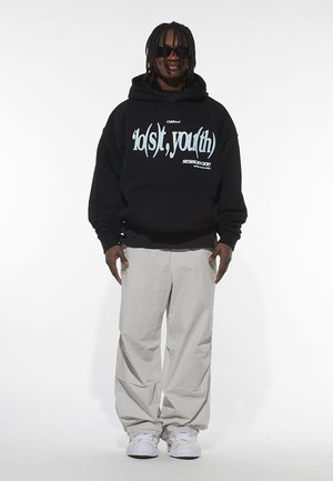 Black hoodie with light blue text, featuring a front pocket and drawstring hood, paired with light gray baggy pants and white sneakers.
