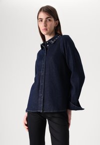 Camicia in denim blu scuro con maniche lunghe e collo a barchetta. Presenta bottoni metallici sul collo e una vestibilità dritta.