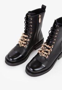 Stivali ankle in pelle nera con punta lucida, dettagli in hardware color oro e perline decorative lungo i lacci.
