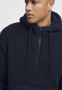 Marineblå hettegenser i fleece med en strukturert, plyssete overflate, med en halv glidelås, justerbar snor i hetta og frontlomme.