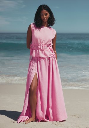 Femme debout pieds nus sur une plage de sable, portant un haut rose sans manches et une jupe longue assortie avec une fente haute, océan et ciel en arrière-plan.