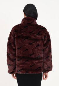 Kvinna med svart hår iförd en burgundyröd fuskpälsjacka med svarta Nike-logotyper, sedd bakifrån mot en vit bakgrund.
