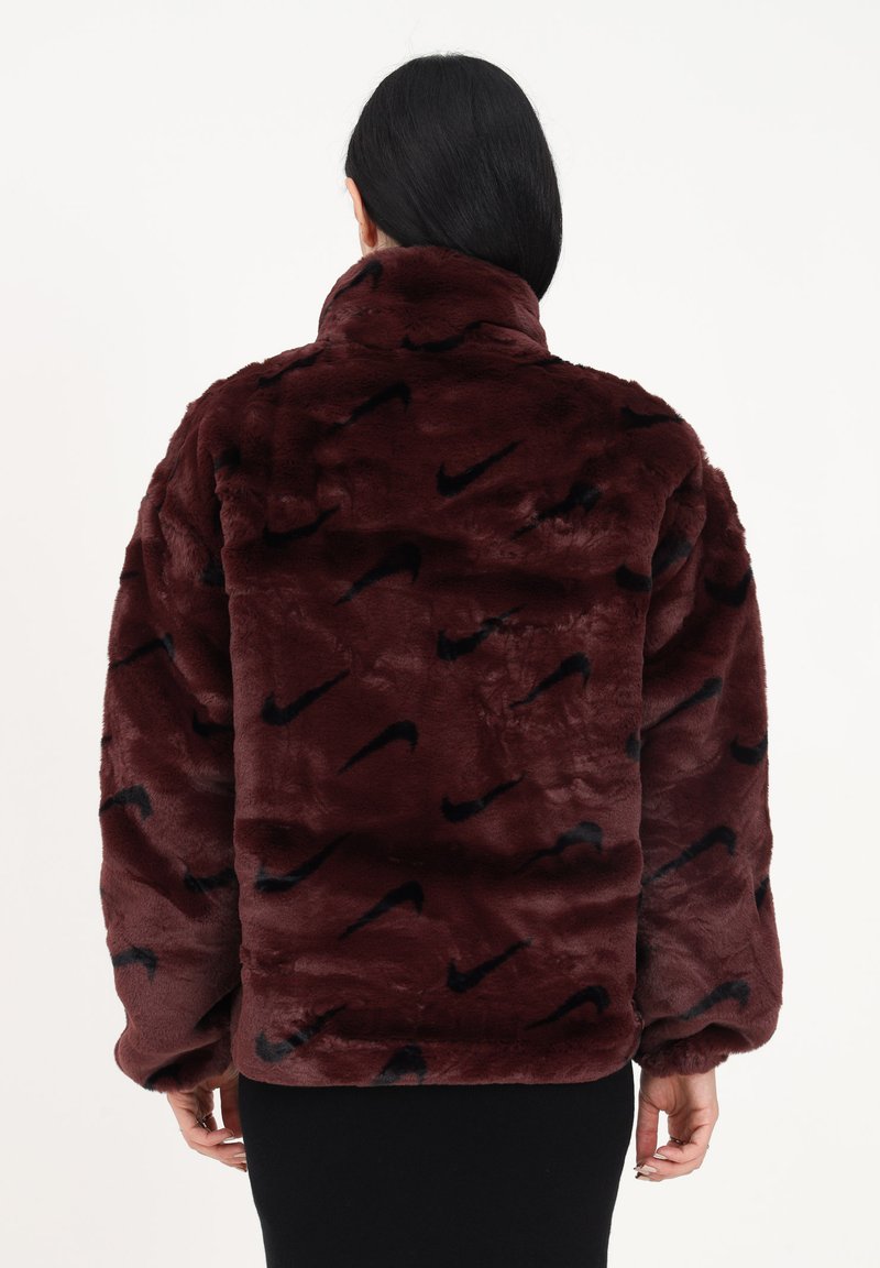 Femme aux cheveux noirs portant une veste en fausse fourrure bordeaux avec des logos Nike noirs, vue de dos sur un fond blanc.
