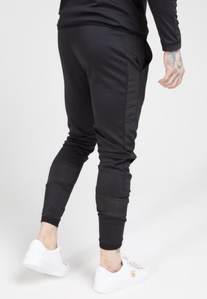 Pantalons de jogging noirs au fit ajusté, à la texture lisse, avec des poches latérales et des accents de panneaux contrastants. Assortis à des baskets blanches.