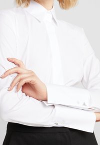 Person in einem weißen Langarmhemd mit Manschettenknöpfen, verschränkten Armen und schwarzen Hosen, die bis zur Taille sichtbar sind.