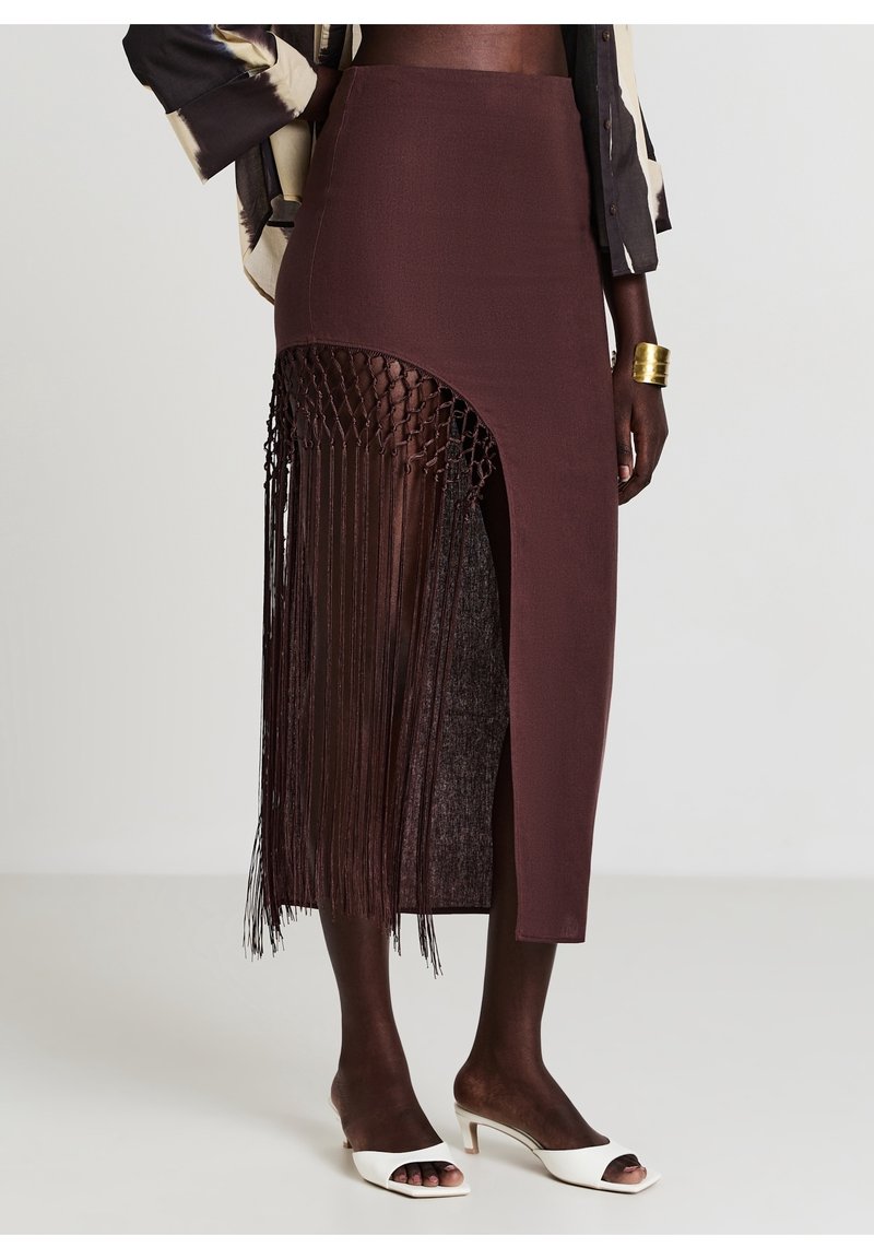Femme portant une jupe marron taille haute avec des franges en macramé sur un côté et des sandales blanches à talons ouverts.