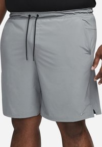 Homme portant un short de sport gris avec des cordons noirs, des poches latérales et un petit logo Nike près de l’ourlet sur la jambe droite.