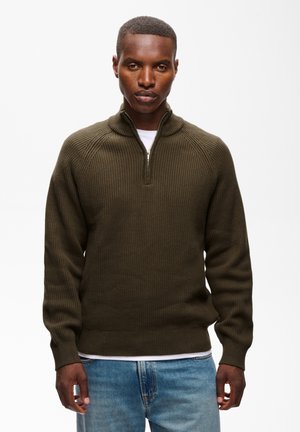Homme portant un pull zippé côtelé vert olive à manches longues et un jean bleu, debout devant un fond blanc.