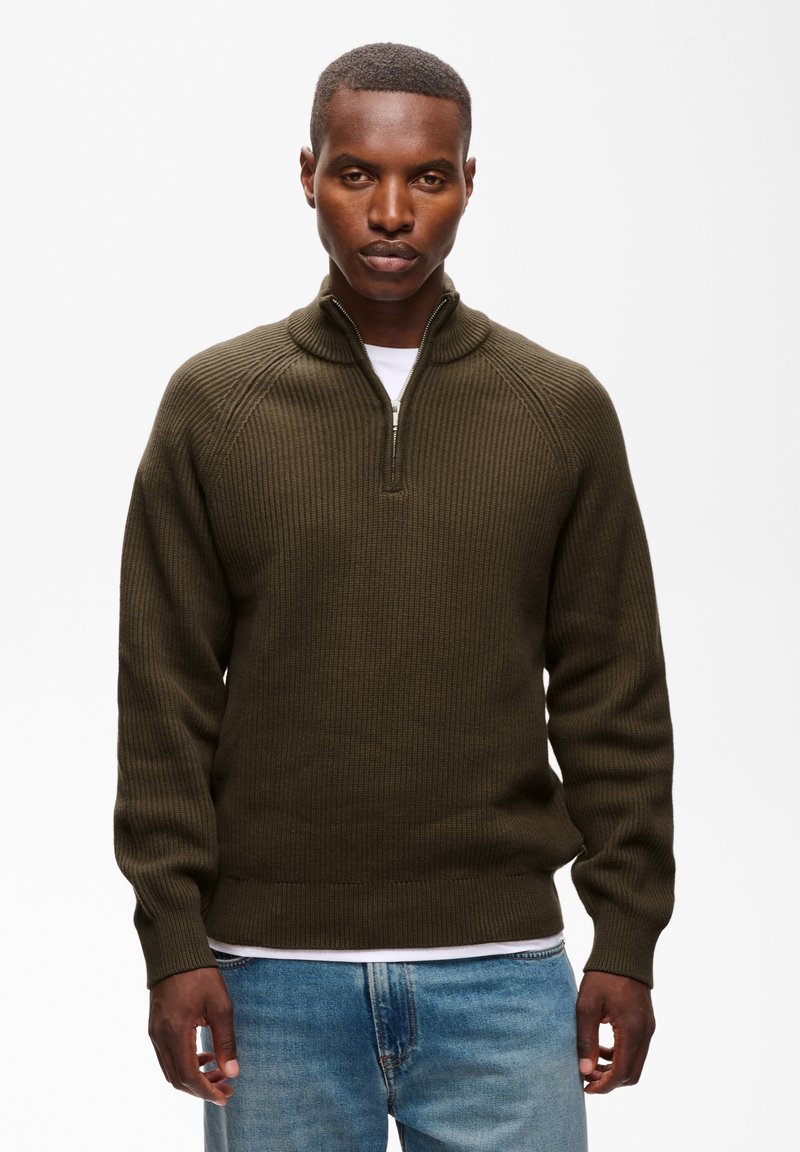Homme portant un pull zippé côtelé vert olive à manches longues et un jean bleu, debout devant un fond blanc.