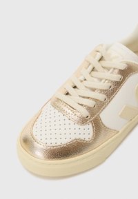 Sneaker bianca e oro con una tomaia in sintetico testurizzato, punta perforata, lacci crema e suola in gomma con un motivo sottile.