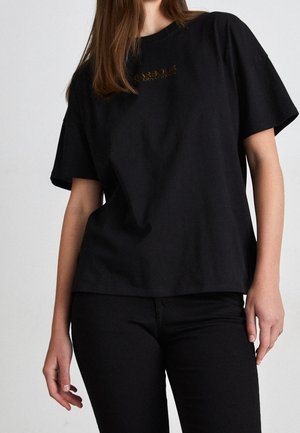 Kobieta nosząca luźną czarną koszulkę z delikatnym złotym napisem i dopasowane czarne spodnie, stojąca na tle jednolitego jasnego tła.