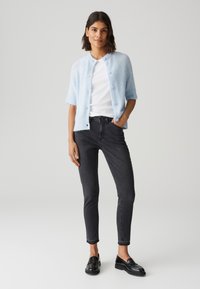 Cardigan en peluche bleu clair avec des manches courtes et un col rond, fermeture par boutons. Porté sur un t-shirt blanc et un jean skinny noir. Mocassins noirs.