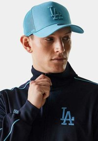 Gorra de béisbol azul claro con el logotipo 'LA' en relieve, combinada con un chándal oscuro. La gorra tiene la parte trasera de malla y visera curva.