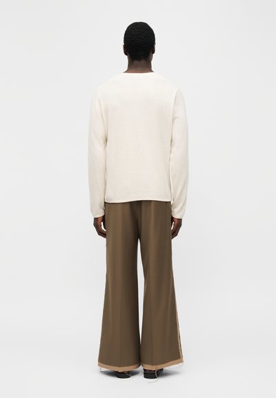 Άνδρας που έχει την πλάτη γυρισμένη, φοράει λευκό πουκάμισο με μακριά μανίκια και καφέ παντελόνι φαρδύ στο κάτω μέρος, μπροστά σε απλό λευκό φόντο.