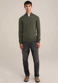 Suéter verde con cuello con cremallera, hecho de una tela suave. Combinado con jeans gris oscuro y botas marrón claro, de pie sobre un fondo neutro.
