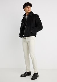 Νεαρός άνδρας που φοράει μαύρο τζάκετ με φερμουάρ, ανοιχτό μπεζ παντελόνι και μαύρα μοκασίνια, στέκεται με τα χέρια στις τσέπες του τζάκετ, κοιτάζοντας προς το πλάι.