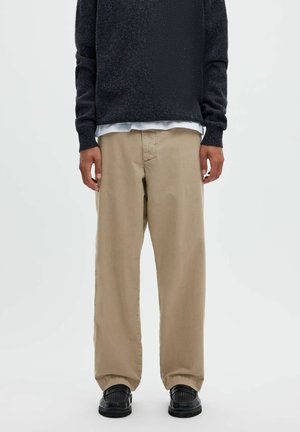 Personne portant un pantalon beige ample, des chaussures noires texturées, un pull sombre et une chemise blanche en dessous, debout devant un fond uni.