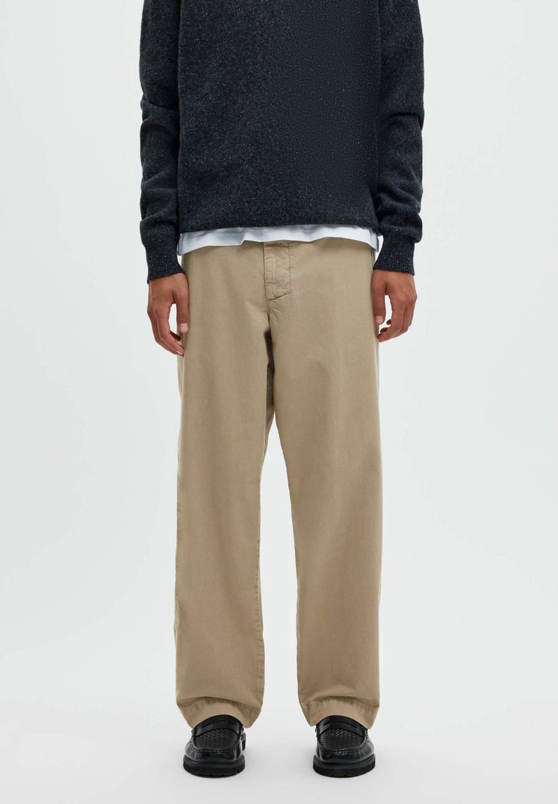 Personne portant un pantalon beige ample, des chaussures noires texturées, un pull sombre et une chemise blanche en dessous, debout devant un fond uni.
