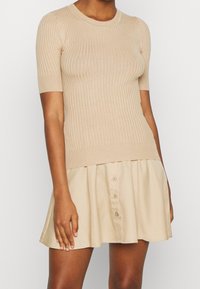 Beige ribbad stickad topp med korta ärmar, rund halsringning och en figurnära midja, som är ihop med en svängig beige knappad kjol. Mjuk textur.