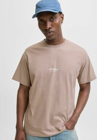 Tricou cu mânecă scurtă, de culoare maro deschis, cu guler rotund și logo-ul alb "JJ Studios" imprimat pe piept; purtat cu blugi de denim decolorați.