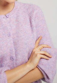 Cardigan en lavande avec un tricot texturé, comportant des manches courtes, un col rond, et deux boutons violets à l'avant.