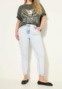 Šedé grafické tričko s designem orla a textem "BUĎ ODVAŽNÝ A DIVOKÝ", světle modré úzké džíny a černé ploché boty.