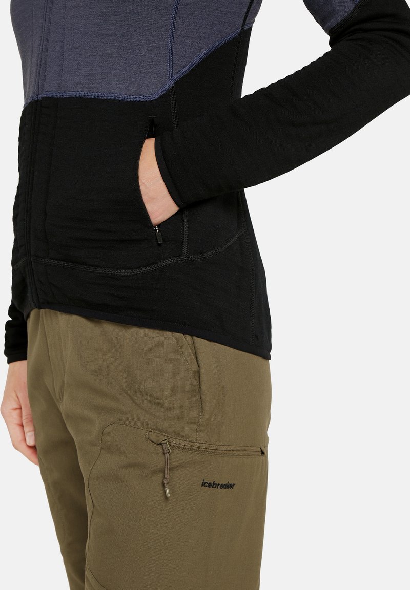 Schwarzes und graues Langarmoberteil aus strukturiertem Stoff mit halbem Reißverschluss und Seitentasche. Kombiniert mit olivgrünen Hosen mit Reißverschlusstasche.