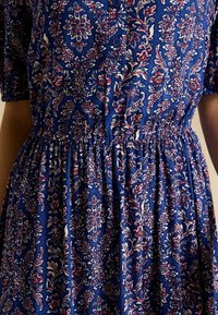 Modrá kvetinová šaty s button-down predným zapínaním, krátkymi rukávmi, zberaným pásom a zložitými bielymi, červenými a ružovými vzormi na textúrovanej látke.