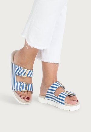 Füße, die blau-weiß gestreifte Sandalen mit goldenen Schnallen tragen, kombiniert mit weißen, kurz geschnittenen, ausgefransten Hosen vor hellem Hintergrund.