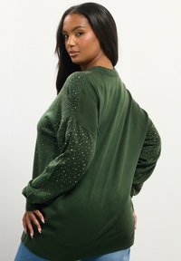 Frau mit langen, dunklen Haaren trägt einen grünen Pullover mit kleinen weißen Punkten auf den Ärmeln und posiert mit den Händen in den Hüften vor einem einfachen hellen Hintergrund.