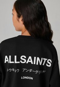 Juodas medvilnės džemperis su ryškiu baltu tekstu nugarinėje pusėje, kuriame parašyta "ALLSAINTS" anglų ir japonų kalbomis, o po juo yra užrašas "LONDON".
