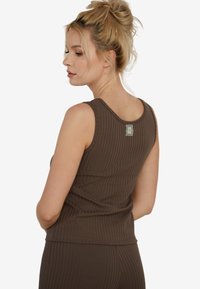 Blondynka z włosami związanymi w kok, ubrana w bezrękawnik z brązowej, prążkowanej tkaniny, pokazana od tyłu na białym tle.