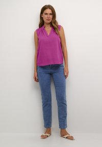 Femme se tenant contre un mur blanc, portant un haut sans manches magenta, un jean bleu et des sandales beiges.
