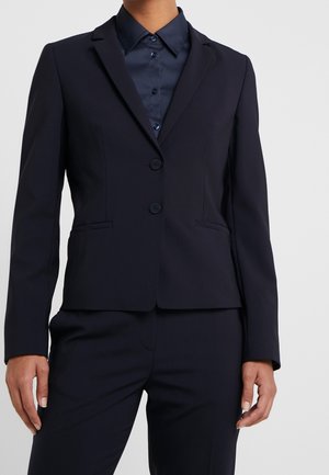 Person iført en tætsiddende mørk marineblå blazer med to knapper over en matchende mørk marineblå skjorte med knapper og matchende bukser.