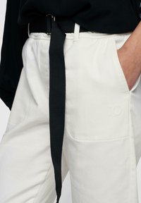 Pantaloni bianchi di cotone con una cintura intrecciata nera. Presentano due tasche frontali e un dettaglio stampato "10" sul lato sinistro. Tessuto morbido e vestibilità rilassata.