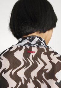 Persona con capelli neri corti che indossa una camicia trasparente a motivi con forme nere, bianche e un logo rosso 'HUGO' sul colletto posteriore.