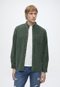 Joven con cabello rubio que lleva una camisa verde abotonada sobre una camiseta blanca y vaqueros azules rotos, de pie contra un fondo claro y liso.