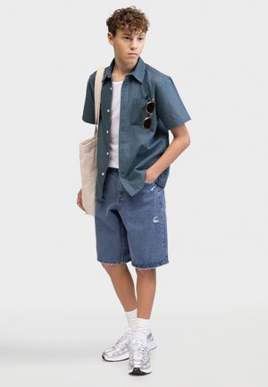 Νεαρό άτομο που φοράει μπλε καρό κοντομάνικο πουκάμισο, λευκό t-shirt, τζιν σορτς, λευκές κάλτσες, ασημένια αθλητικά παπούτσια, κουβαλώντας μπεζ τσάντα tote.