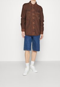 Brun flanellskjorta med två bröstfickor och knappstängning, kombinerad med blå denimshorts och vita sneakers, stiliserad med vita strumpor.