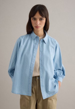 Junge Frau mit langen braunen Haaren, die ein blau-weiß gestreiftes Hemd über einem beigen Top und khakifarbene Hosen trägt, mit den Händen in den Taschen.