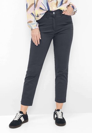 Kvinde iført sorte tætsiddende bukser, farverig bluse med abstrakt mønster og sorte sneakers med hvide snørebånd, stående mod en hvid baggrund.