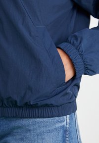 Casaco corta-vento azul marinho com punhos e cintura elásticos. Apresenta um bolso frontal e um tecido leve e texturizado.