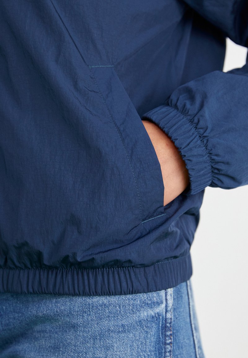 Coupe-vent bleu marine avec des poignets et une taille élastiques. Présente une poche avant et un tissu léger et texturé.