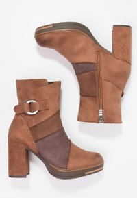 Des bottines en suede marron présentent un talon haut, une fermeture éclair sur le côté et un design en blocs de couleurs avec des accents marron foncé et un détail de courroie circulaire.