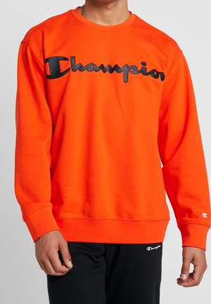 Man draagt een fel oranje lange mouwen sweatshirt met een groot zwart 'Champion' logo op de borst en een kleiner logo op de linker mouwboord.