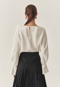 Biała bluzka z długim rękawem i okrągłym dekoltem, z tyłu z wycięciem w kształcie klucza i falbaniastym detalem, zestawiona z czarną plisowaną spódnicą o wysokim stanie.